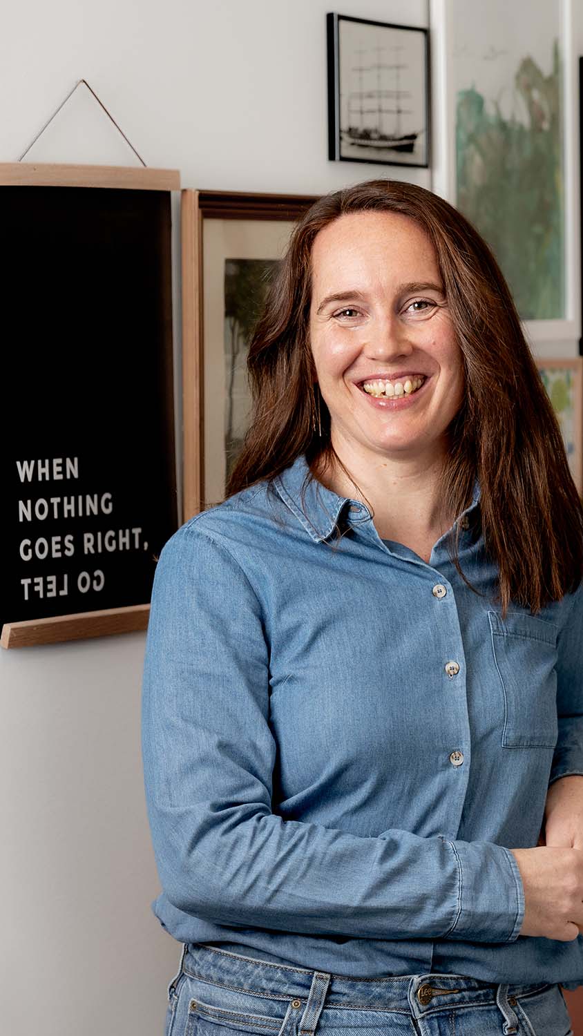 Ett foto på en leende Anna-Karin Mikkelä som står i blå skjorta lutad mot ett piano. På väggen bakom henne syns en tavla: "When nothing goes right, go left." Foto av Jessica Rankell
